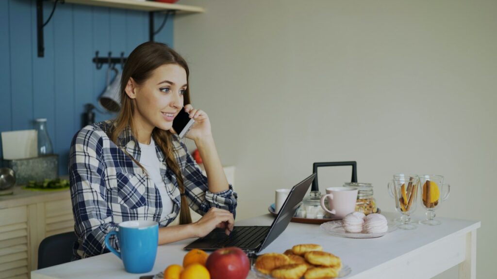 Woman on a phone call while working on a laptop in her kitchen