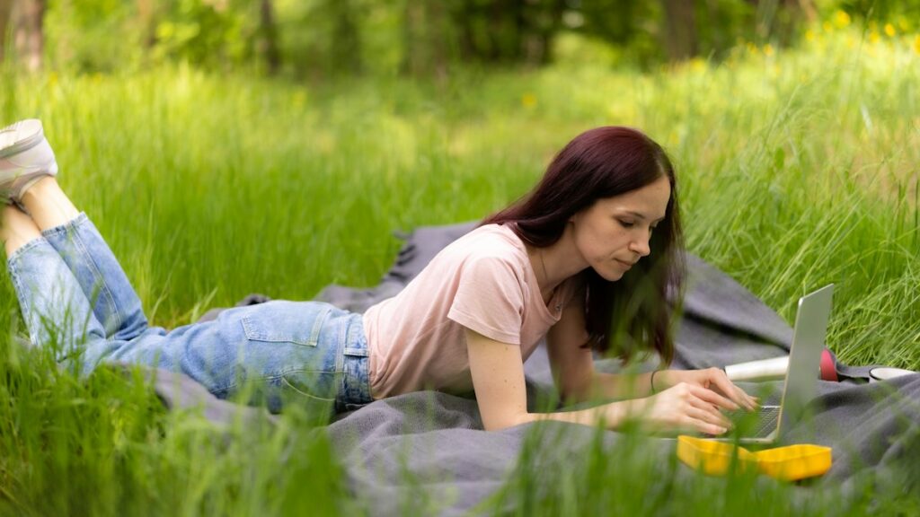 Woman working on a laptop while lying on a blanket in a park
